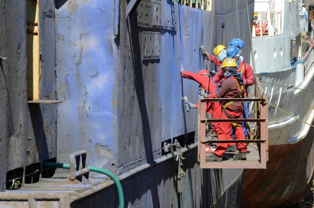 img-09-free-img.jpg Workers in safety gear repairing a ship's hull at a busy shipyard.